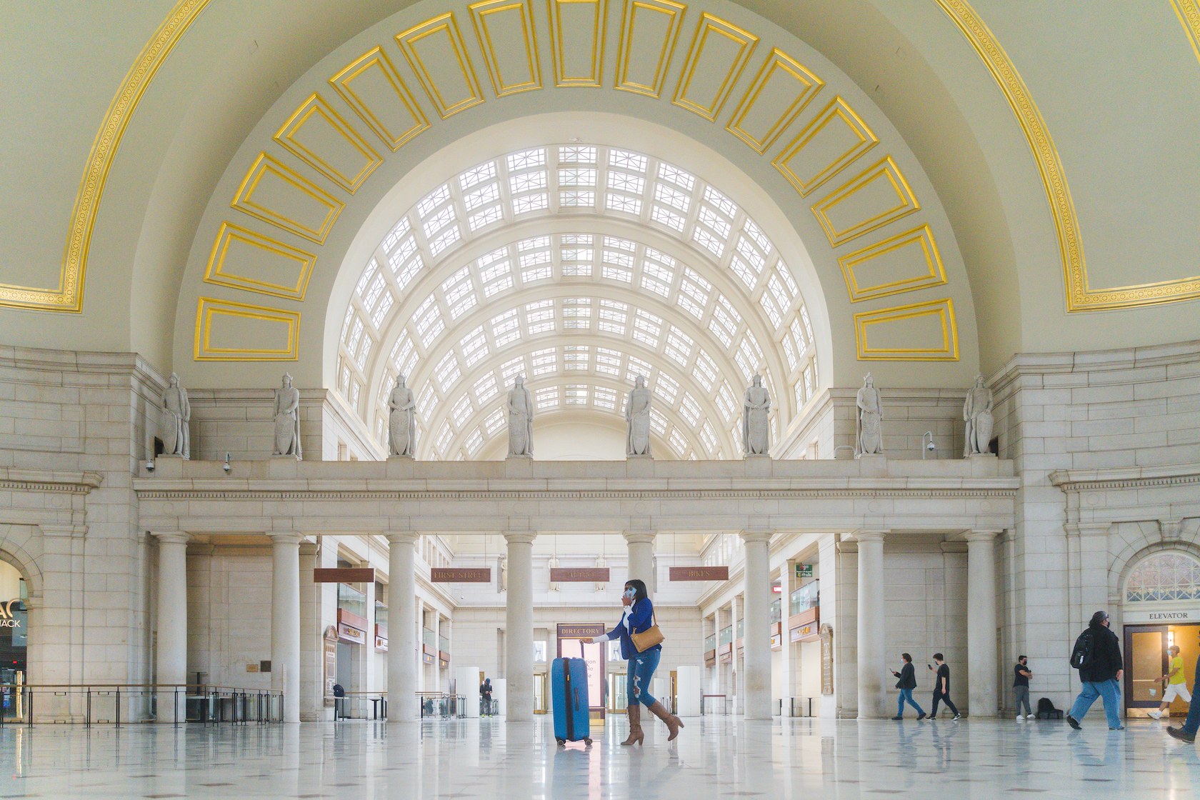  A woman in a blue coat and brown boots walks with a blue suitcase through the grand, arched interior of a spacious train station adorned with statues and large windows.